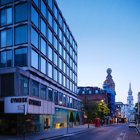 Hotel St Martins Lane Londra Esterno foto The photo shows a street scene during twilight, featuring a modern, glass-fronted building on the left side, which appears to be a gym called "GYMBOK GYM." On the right, there are more traditional architectural elements, including a tower and a steep