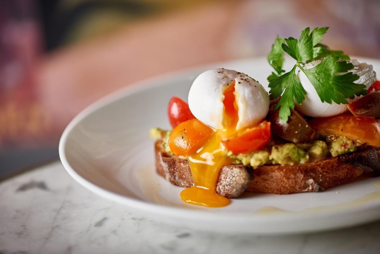 Hotel St Martins Lane Londra Esterno foto The photo showcases a beautifully presented dish on a white plate. It features a slice of brown bread topped with a spread that resembles avocado, accompanied by vibrant cherry tomatoes. Atop the bread, there are two poached eggs, with the yolks runn