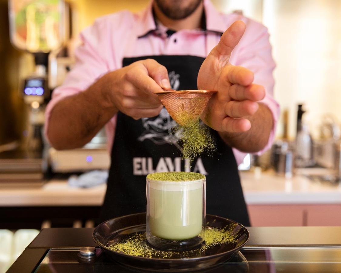Hotel St Martins Lane Londra Esterno foto The image shows a person preparing a drink, likely a matcha-based beverage. They are wearing an apron and using a small scoop to sprinkle what appears to be green matcha powder into a glass. The glass is filled with a light green drink, possibly matc
