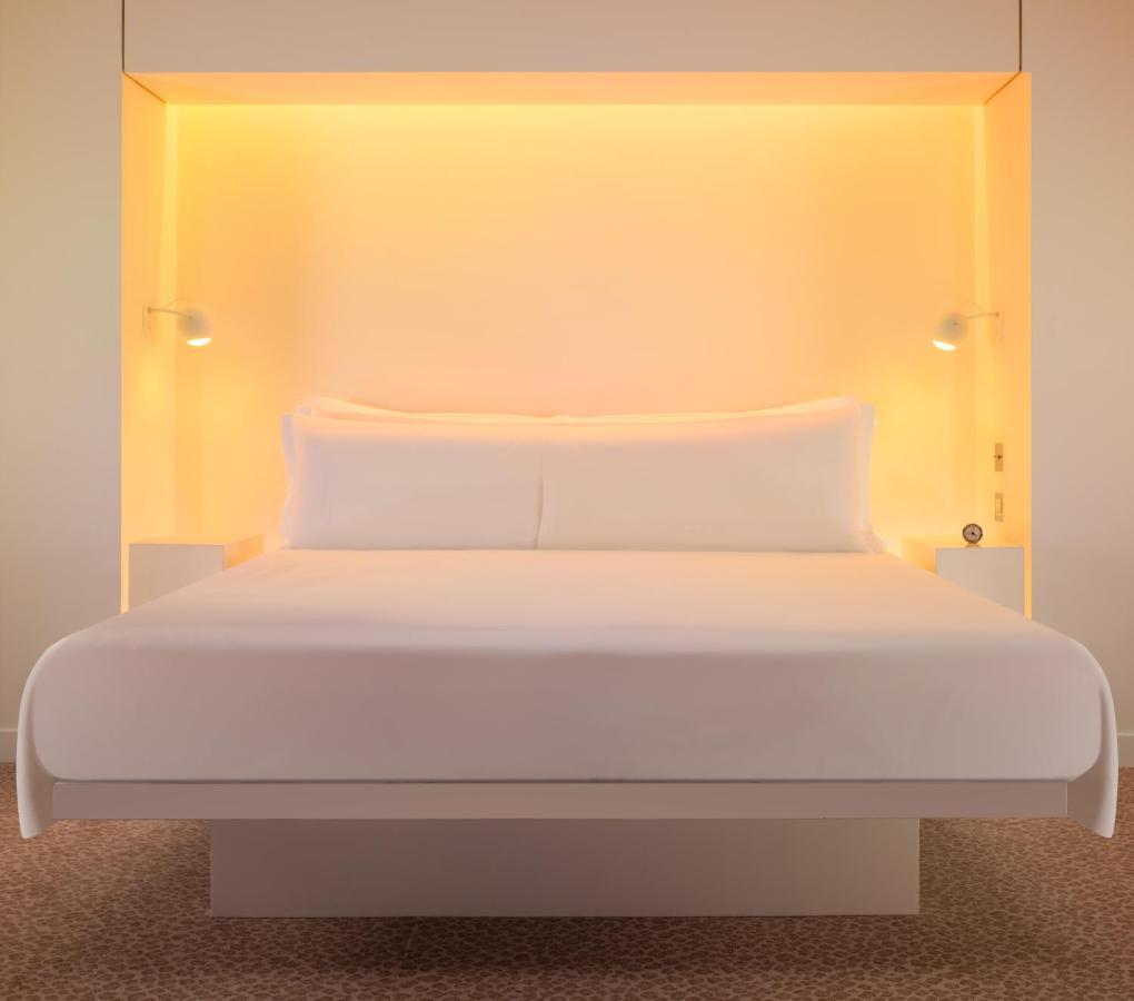 Hotel St Martins Lane Londra Esterno foto The photo shows a modern and minimalist bedroom setup featuring a large white bed with two pillows. The bed is positioned against a glowing orange backdrop, which adds warmth to the ambiance. On either side of the bed, there are small nightstands equ