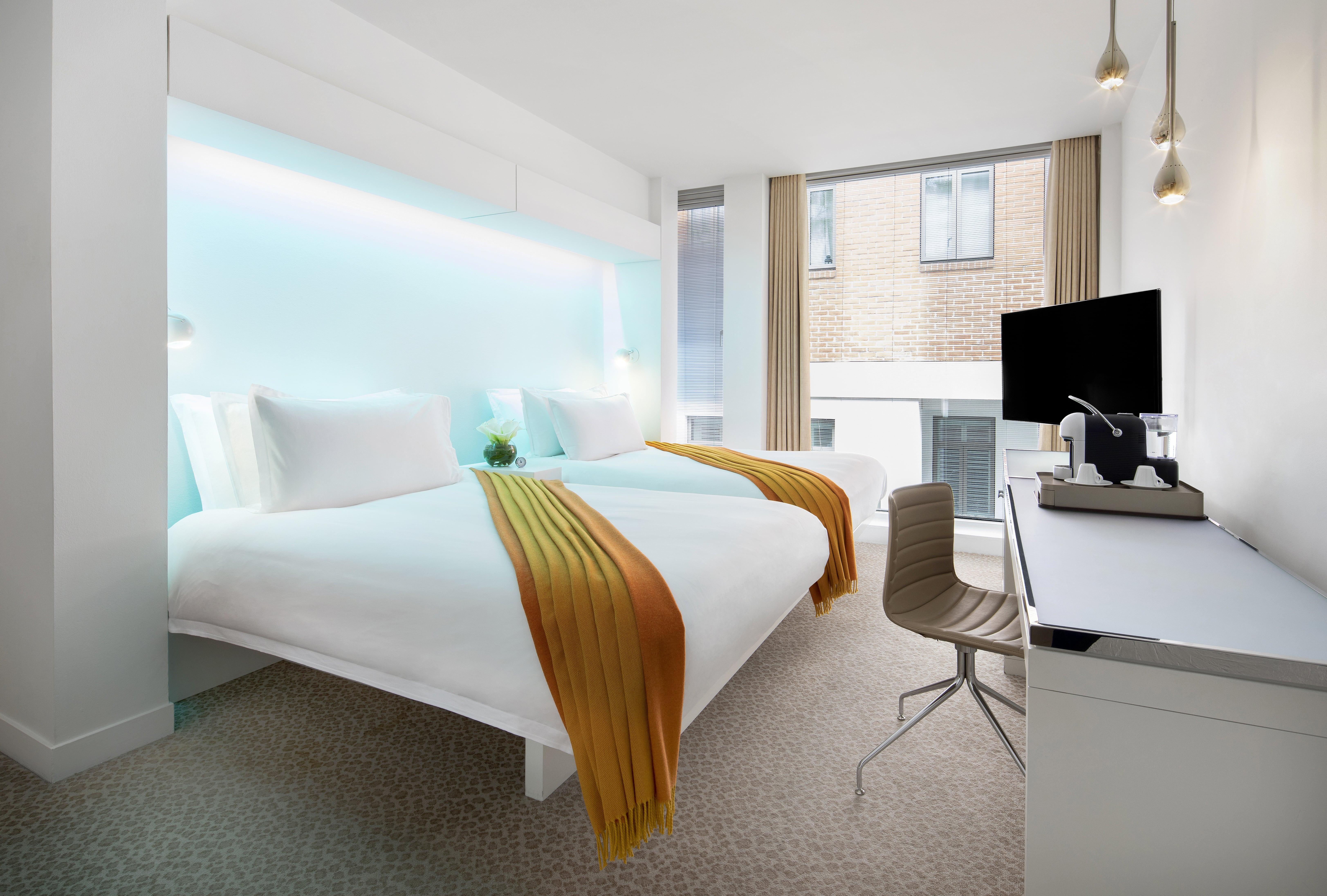 Hotel St Martins Lane Londra Esterno foto The photo shows a modern hotel room with a clean, contemporary design. There are two beds, each with white bedding and vibrant orange and yellow throws. Behind the beds, there is a glowing blue accent wall, adding a stylish touch. The room features a