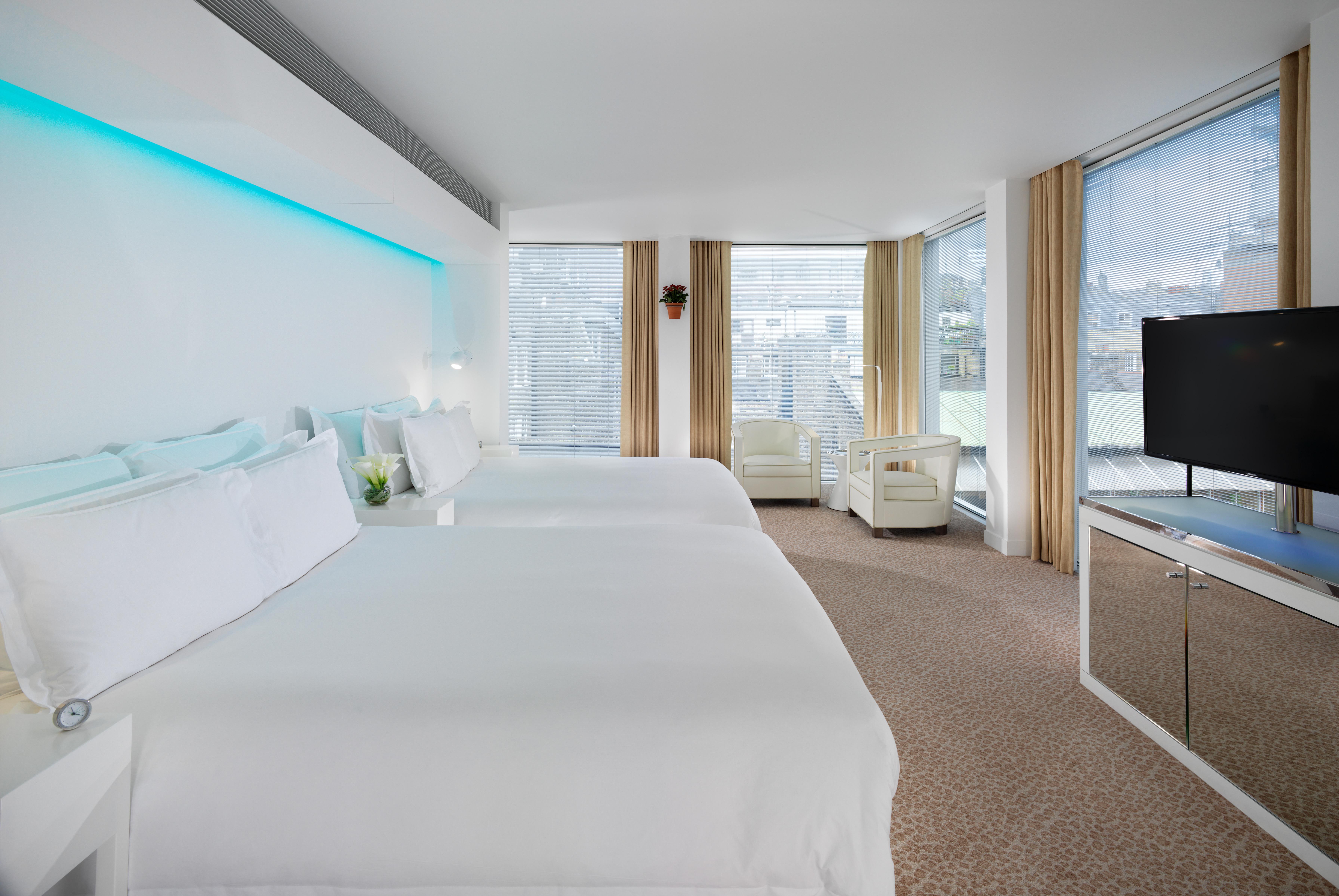 Hotel St Martins Lane Londra Esterno foto The photo shows a modern hotel room featuring two large beds with white bedding. The room has large windows with light-colored curtains, allowing natural light to fill the space. There is a small seating area with a white sofa and a bedside table. A 