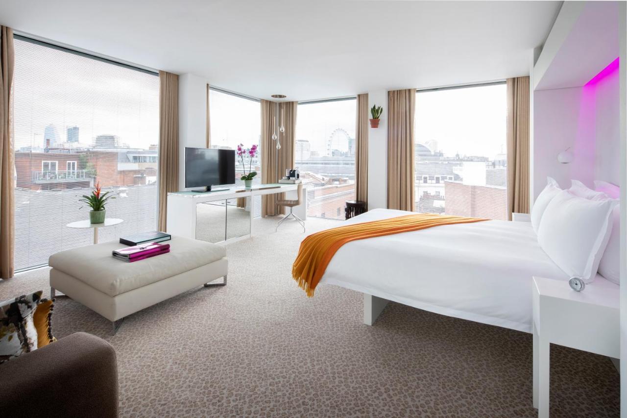 Hotel St Martins Lane Londra Esterno foto The photo shows a modern hotel room featuring a large bed with white linens and an orange throw blanket. The room has ample natural light coming through large windows with beige curtains, offering a view of a cityscape. There is a small seating area 