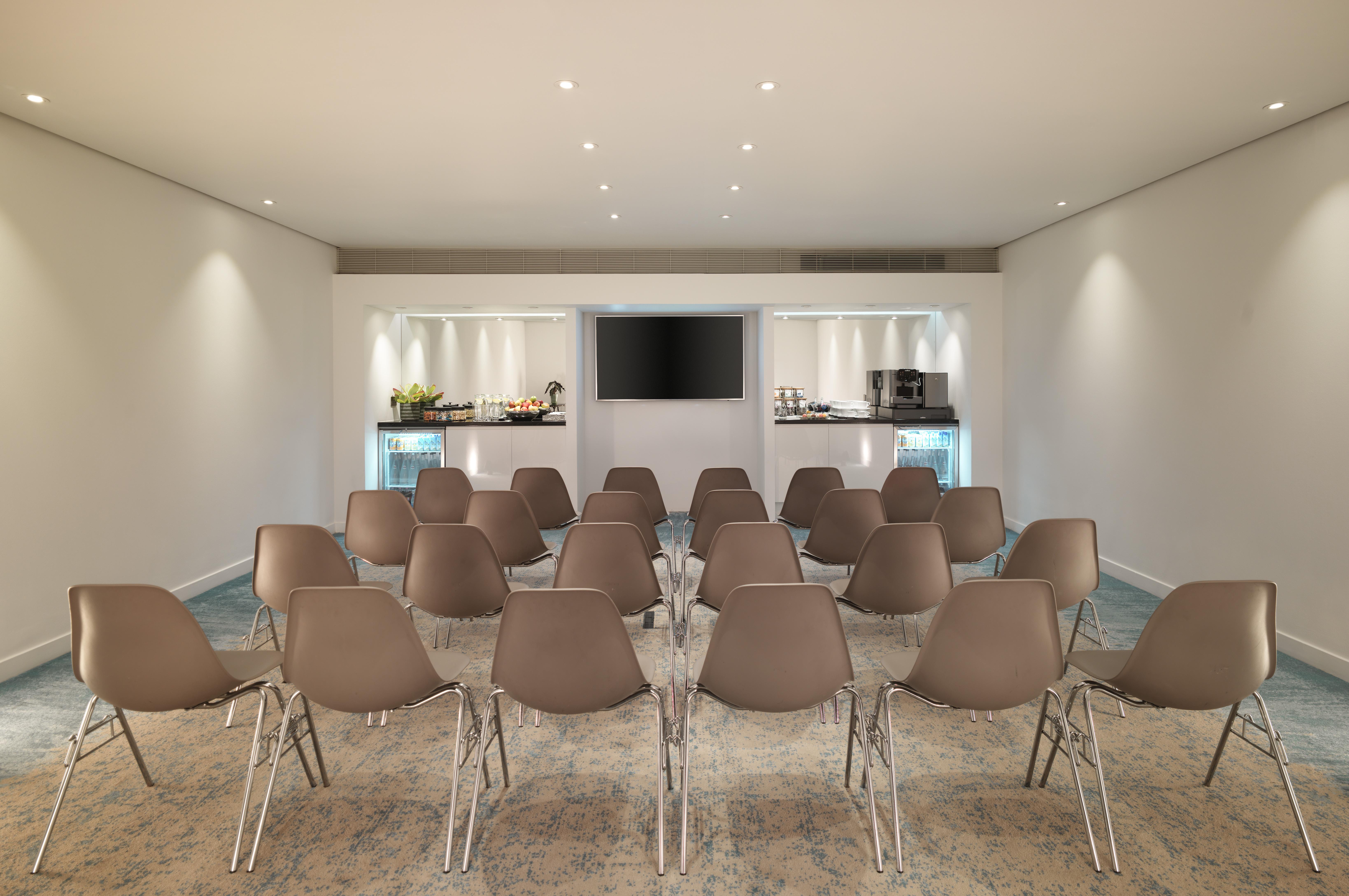 Hotel St Martins Lane Londra Esterno foto The photo depicts a modern meeting room or conference space. The room features several rows of light brown chairs arranged facing a wall-mounted television. In the background, there's a counter with refreshments, likely including food and beverages, 