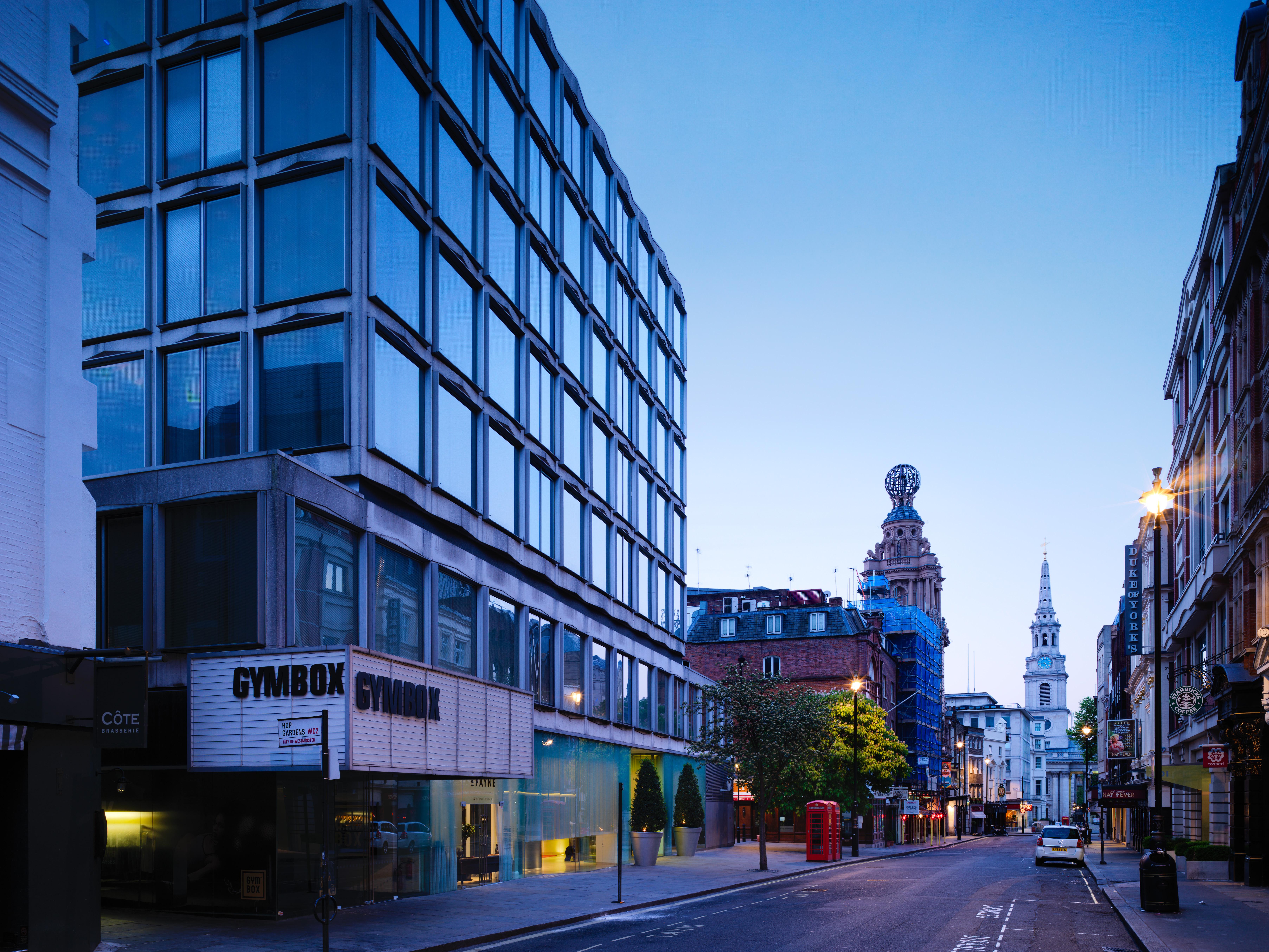 Hotel St Martins Lane Londra Esterno foto The photo shows a street scene during twilight, featuring a modern, glass-fronted building on the left side, which appears to be a gym called "GYMBOK GYM." On the right, there are more traditional architectural elements, including a tower and a steep