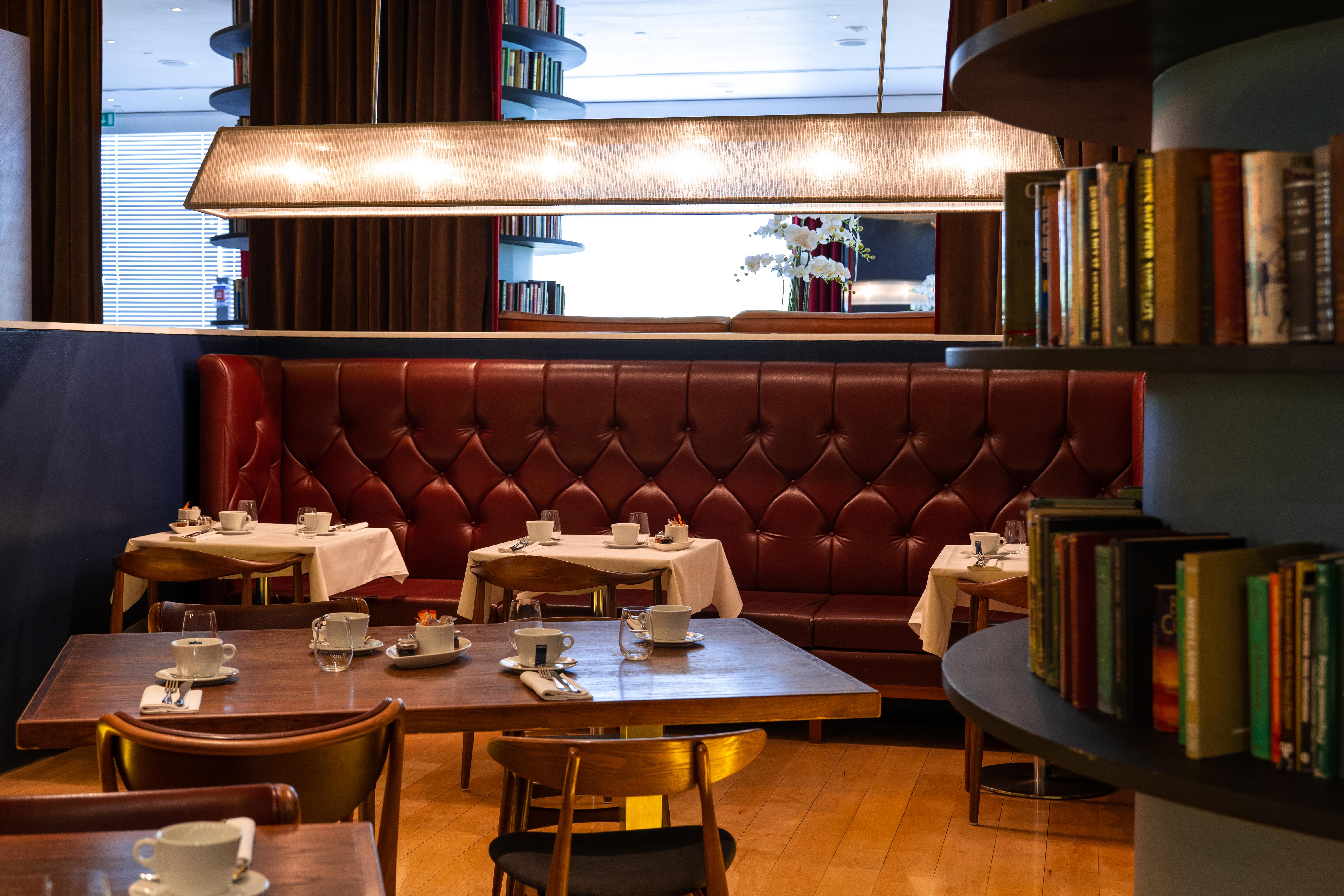 Hotel St Martins Lane Londra Esterno foto The photo depicts a cozy, elegantly designed restaurant interior. There are several round tables set with white tablecloths and accompanied by small white cups, likely for tea or coffee. The seating includes a plush, burgundy leather banquette agains