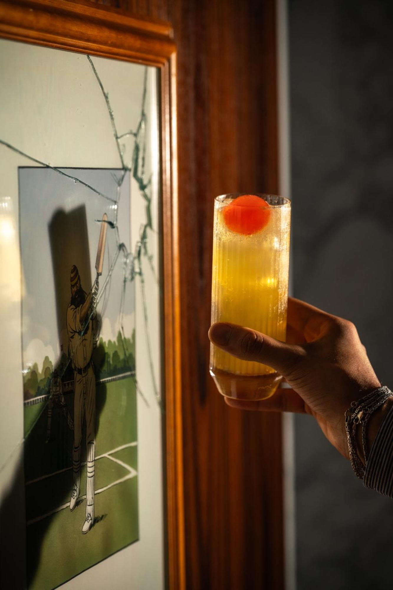 Hotel St Martins Lane Londra Esterno foto The photo shows a hand holding a tall glass containing a drink, which appears to be a yellow cocktail topped with a bright red cherry. In the background, there is a piece of art or a photograph that features a figure, possibly in a baseball or sporti