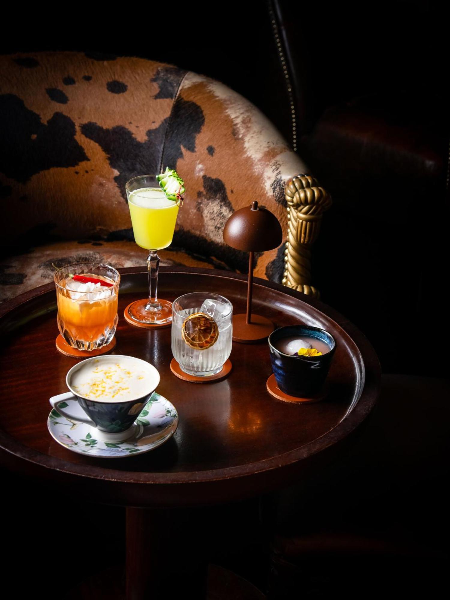 Hotel St Martins Lane Londra Esterno foto The photo displays a variety of drinks arranged on a round wooden tray. There are five distinct beverages:

1. **A light green cocktail** in a tall glass, garnished with a slice of fruit, suggesting a refreshing flavor.
2. **An orange drink** served 