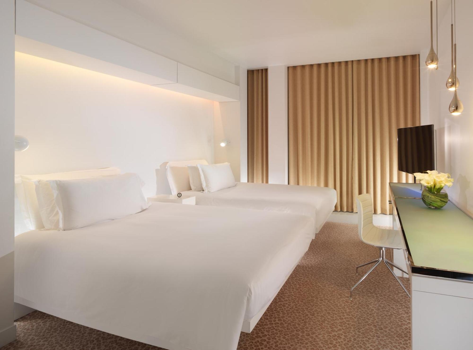 Hotel St Martins Lane Londra Esterno foto The photo shows a modern hotel room featuring two white beds neatly made with minimalistic design. The walls are primarily white with warm lighting highlighting the space. There are large windows dressed with light brown curtains. A small desk with a