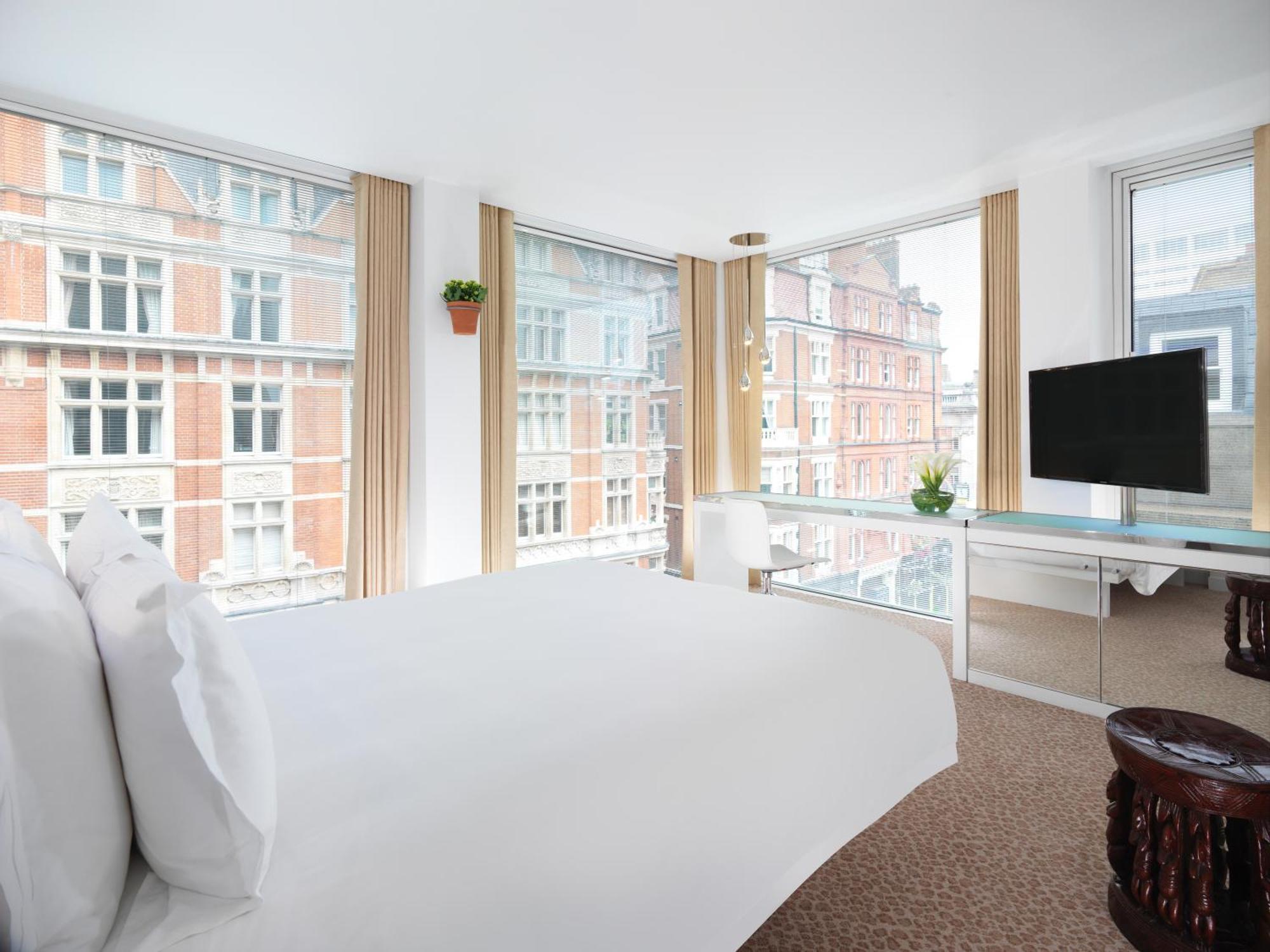 Hotel St Martins Lane Londra Esterno foto The photo shows a modern hotel room featuring a large bed with white linens, positioned near large windows that provide abundant natural light. The windows offer a view of brick buildings outside. There is a flat-screen TV mounted on the wall, a mini