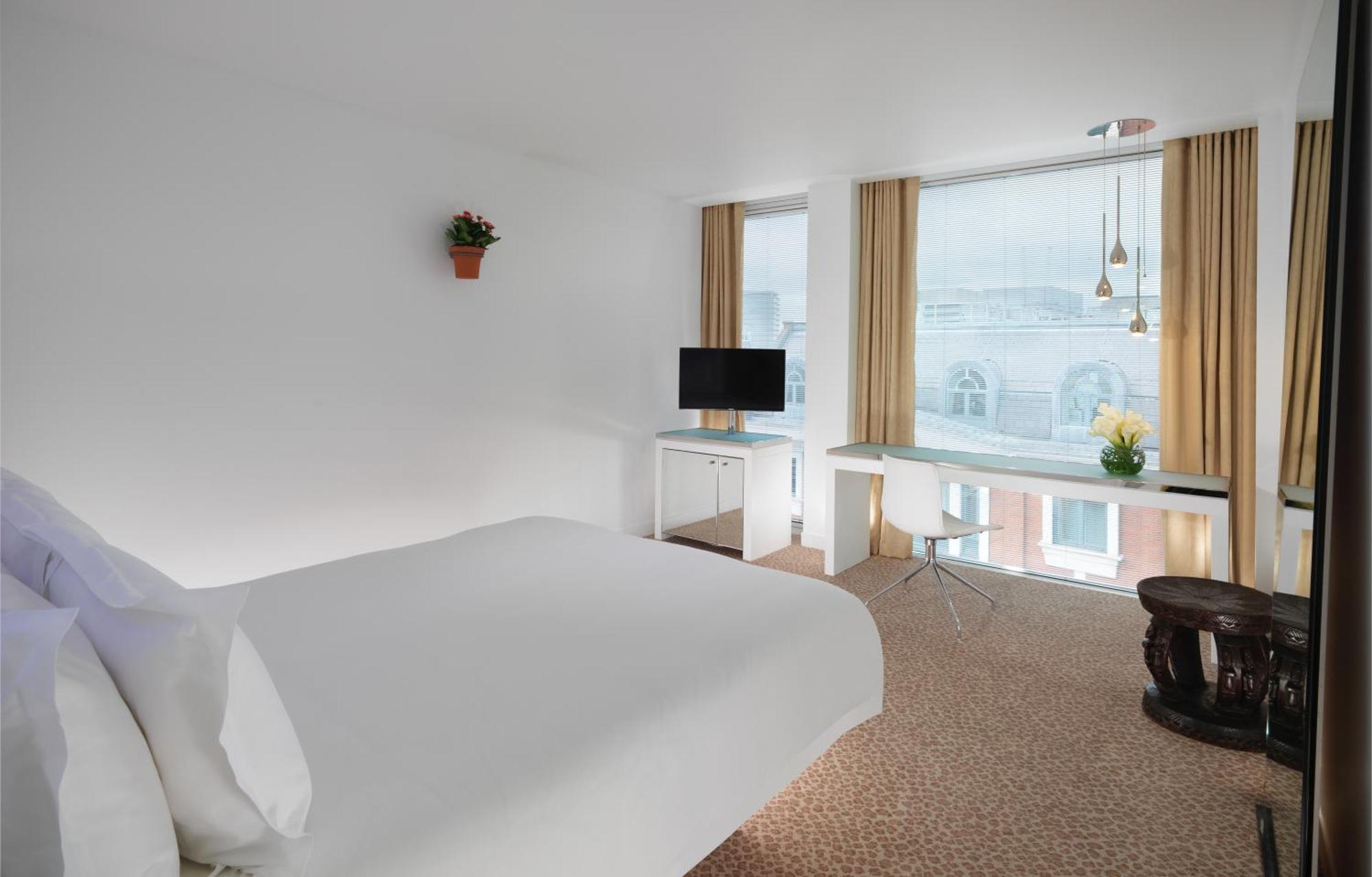 Hotel St Martins Lane Londra Esterno foto The photo shows a modern hotel room that features a large bed with white linens, a small desk with a chair, and a television mounted on the wall. There are large windows covered with light curtains, allowing natural light to fill the room. A small po