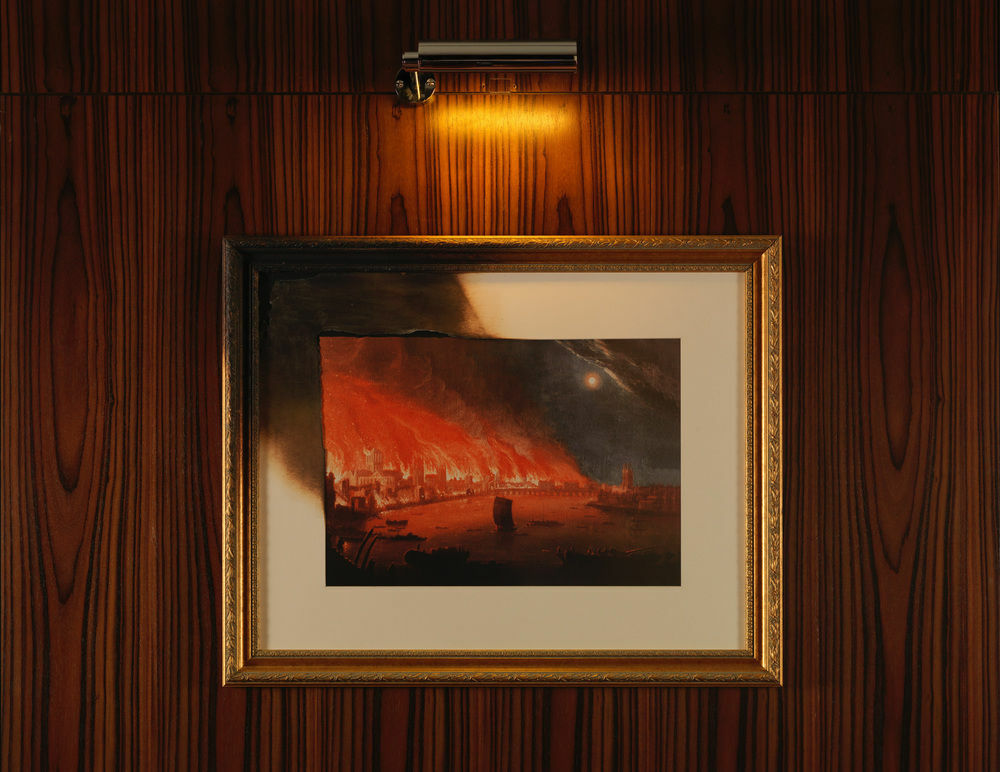 Hotel St Martins Lane Londra Esterno foto The photo shows a framed painting illuminated by a small light above it. The artwork depicts a dramatic scene with fiery red and orange hues, suggesting a sense of destruction or chaos. The painting is set against a wall with a wood grain texture, an